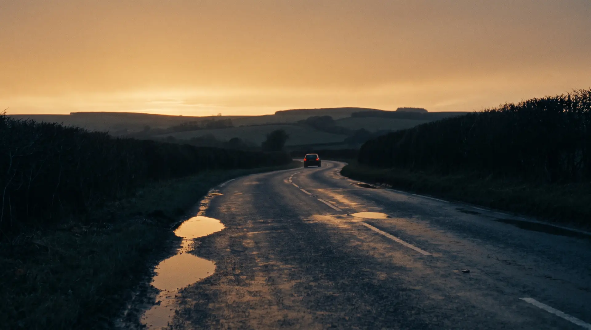 A car driving away into the distance on a wet British A-road at dusk, tail lights lit